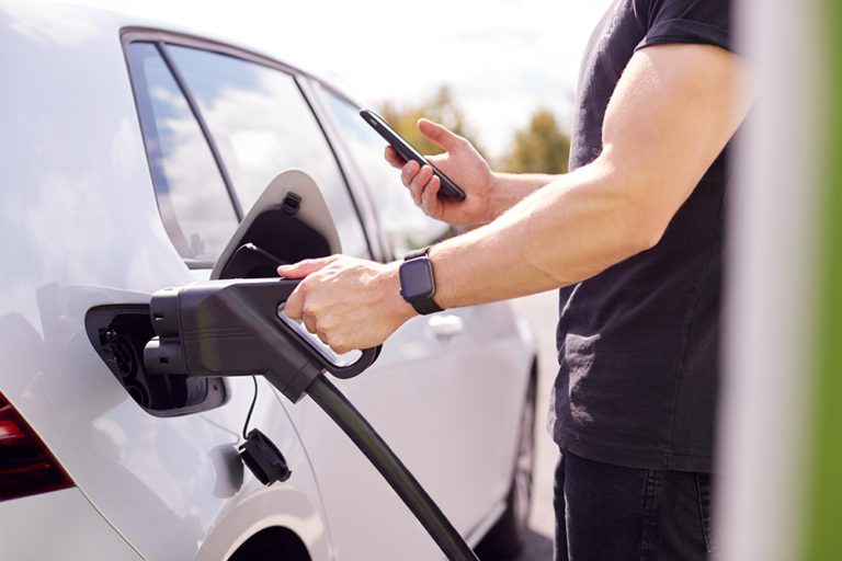 Man Charging Electric Vehicle With Cable Looking At App On Mobile Phone