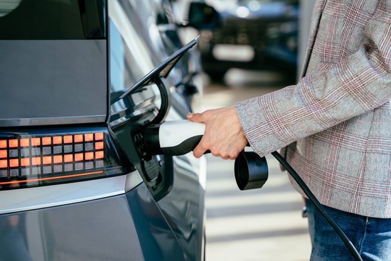 Woman holding charging cable for electric car , looking happy waiting electric car to charge. Caucasian female stands near electric auto in dealership. Smart ecological living